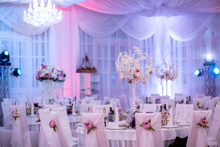 Elegant wedding setup with draped ceiling, floral centerpieces, and ambient lighting. Tables feature detailed place settings.