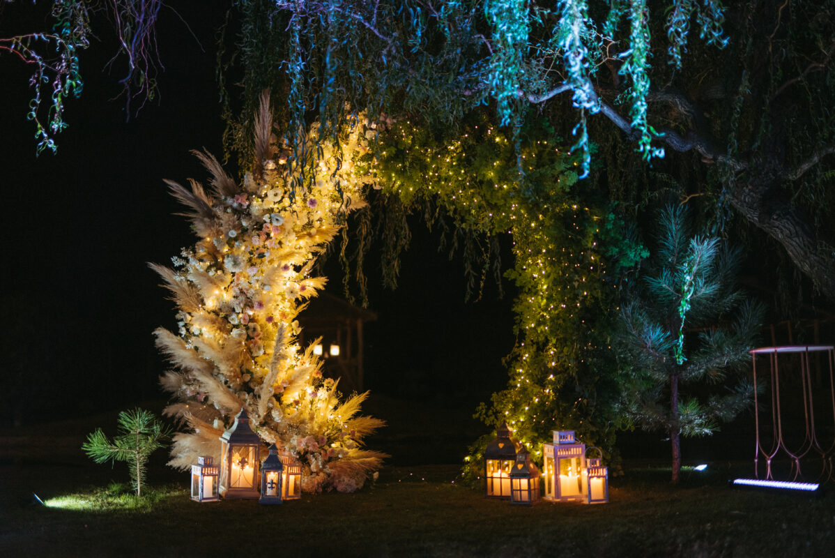 Floral arch with fairy lights and lanterns in an outdoor wedding ceremony setup at night.