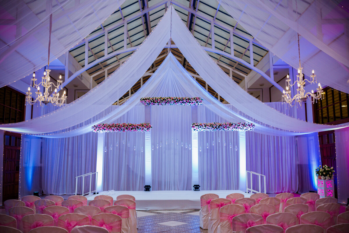Elegant wedding venue setup with draped fabric ceiling, floral arrangements, chandeliers, and seating for a ceremony.