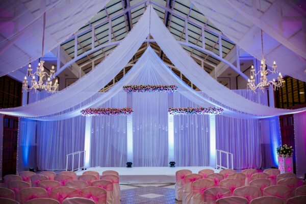 Elegant wedding venue setup with draped fabric ceiling, floral arrangements, chandeliers, and seating for a ceremony.